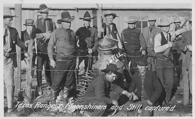 A group of armed Rangers fathered around an alcohol still in front of a barbed wire fence. Writing at the bottom of the photograph reads "Texas Rangers, Moonshiners and Still captured."