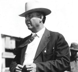A man in a dark suit and tie with a white cowboy hat standing with his hands together.