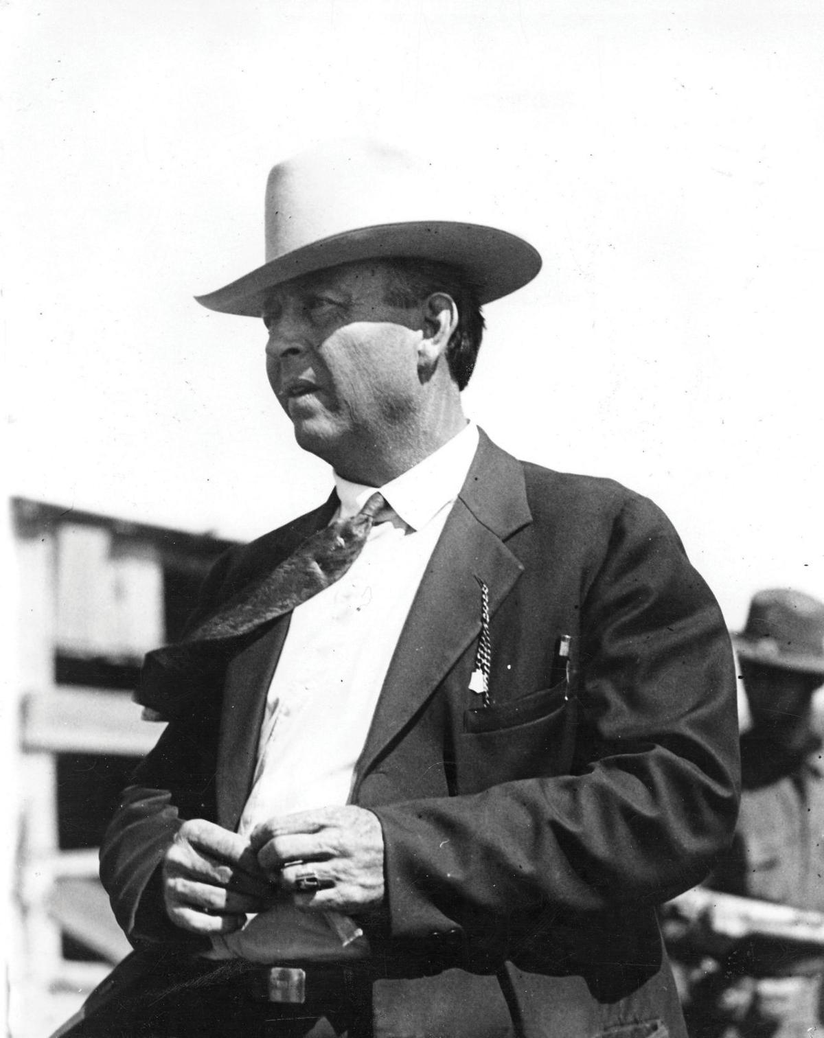 A man in a dark suit and tie with a white cowboy hat standing with his hands together.