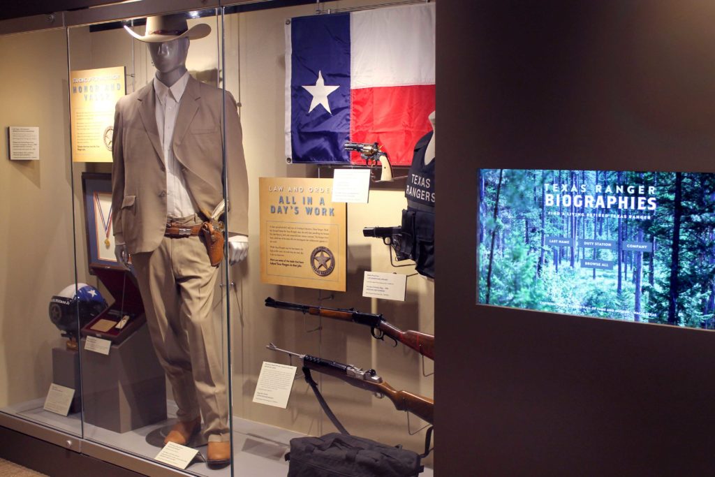 An exhibit with a mannequin wearing a tan suit and cowboy hat, a Texas flag, a flight helmet, four guns, and a bullet proof vest in it. A touch screen with the words "Texas Ranger Biographies" is next to the exhibit.