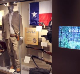 An exhibit with a mannequin wearing a tan suit and cowboy hat, a Texas flag, a flight helmet, four guns, and a bullet proof vest in it. A touch screen with the words "Texas Ranger Biographies" is next to the exhibit.
