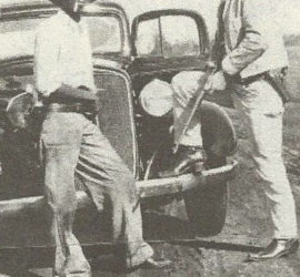 Two men standing in front of a vintage car. One man is holding a firearm.