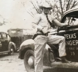 A man in a white hat, light colored pants and button up shirt with tie, and a gun belt and cowboy boots standing next to a car with the words "Texas Rangers Dept. of Public Safety 39" on it.