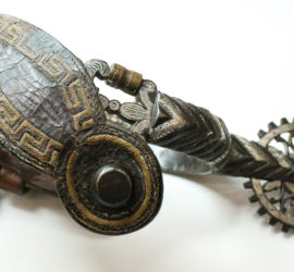 A highly decorative silver boot spur with a dark brown patterned leather strap.