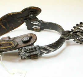 A highly decorative silver boot spur with a dark brown leather strap.