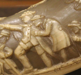 A carving of four men in a shallow boat. One is paddling, two are standing, one is sitting.