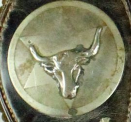 A silver circle engraved with a star and a bull horn attached to dark brown leather with stitching visible.