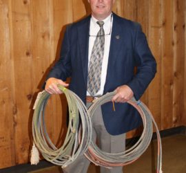 A man in a white hat, dark sport coat, and khaki slacks holds two lariats.