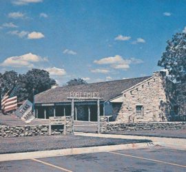 An older photograph with a stone fort style building behind a yard and a short stone wall with three flags: Texas, American, and Texas Ranger. The sign above the entrance gate says "Fort Fisher".