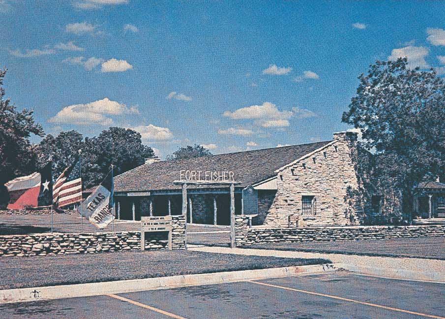 An older photograph with a stone fort style building behind a yard and a short stone wall with three flags: Texas, American, and Texas Ranger. The sign above the entrance gate says "Fort Fisher".