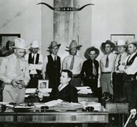 A large group of Rangers in dress shirts standing around a man in a suit seated at a desk in an office.