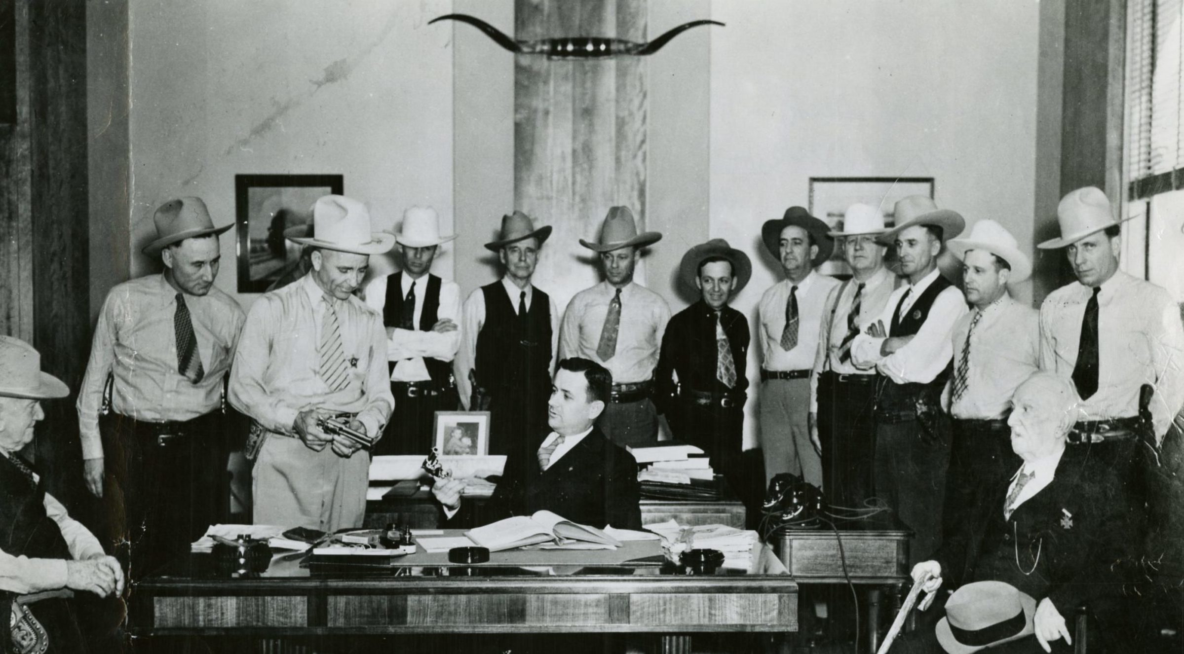 A large group of Rangers in dress shirts standing around a man in a suit seated at a desk in an office.