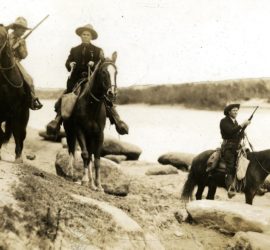 Three armed men in cowboy boots and suits on horseback riding along a riverbank.