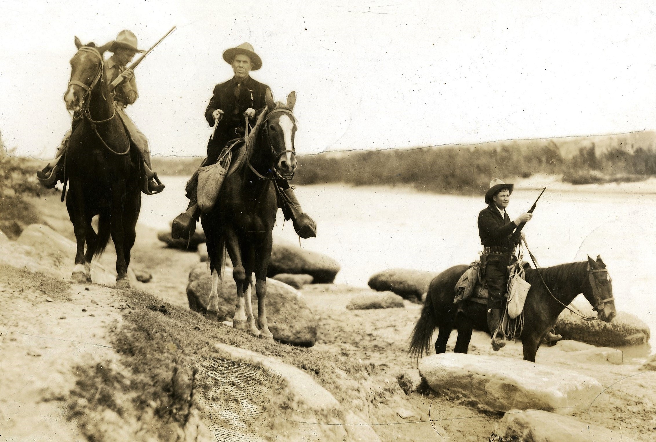 Three armed men in cowboy boots and suits on horseback riding along a riverbank.