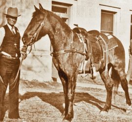 A man in a dark vest and pants over a white shirt with a cowboy hat on stands outside of a building holding the lead rope of a horse. Two other men in suits are in the background.