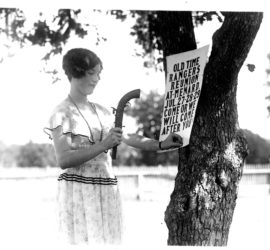 photograph of lady in white dress using gun to hammer sign to tree