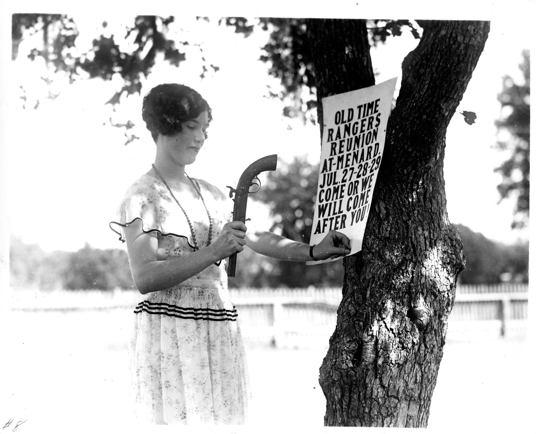 photograph of lady in white dress using gun to hammer sign to tree